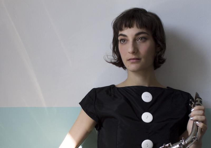 Lea Bertucci is leaning against a wall looking off into the distance holding an alto saxophone