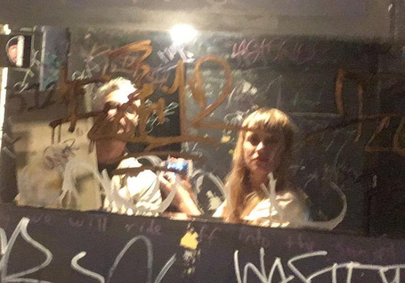 two people stand in front of a dirty graffiti-covered venue bathroom mirror taking a selfie