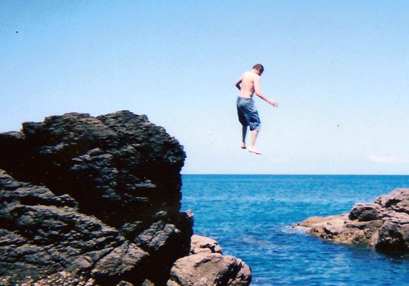 Stefan is floating in mid air. The sky is blue and clear. Srafan  is falling fast into the sea. Stefan is doing a BOMB off the rocks.
