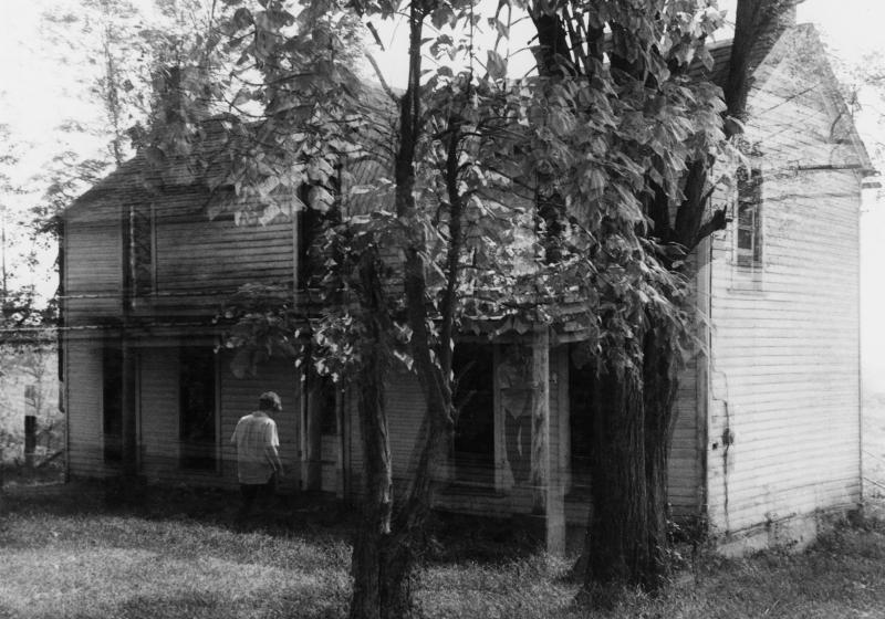 a black and white photo of an old house. The exposure of the photo was clearly long, the house is doubled and ghostly figures stand in various positions in the photo. Photo by Eugene Meatyard