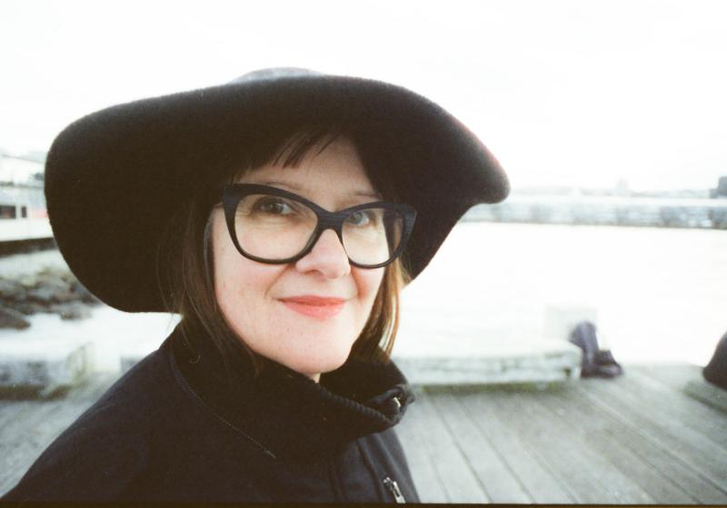 Sonya Waters smiles at the camera in a black hat and black outfit on a pier on the sea