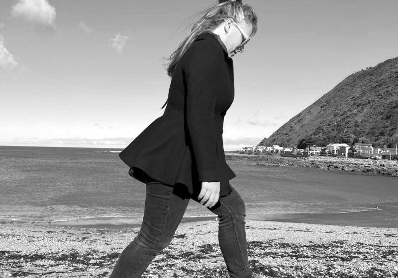 A black and white photo of Rosie Whinray. She walks carefully, one foot at a time along the rocks of a stoney beach. There is a hill cascading down into the sea on the right, off into the distance. The photo is likely taken along the south coast in Wellington, where Rosie collected much of the material for her Pyramid Club exhibition. It is a bright day with few, small clouds. 