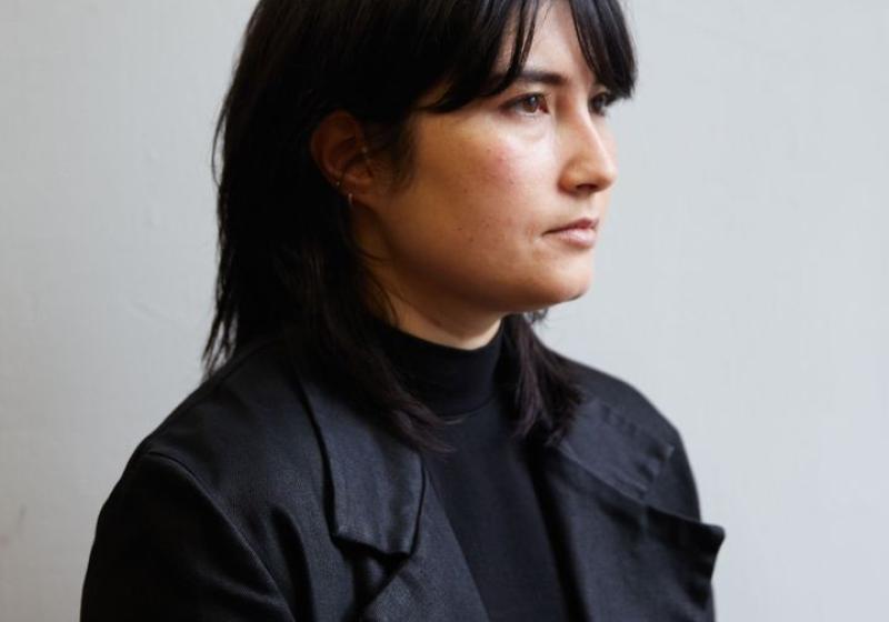 Emi Pogoni sits in front of a grey wall. They are wearing all black and the light is softly catching their face. 