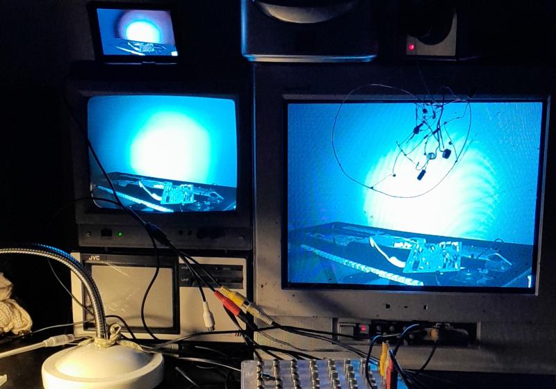 A stack of junk CRT screens stands behind a small musical keyboard with it's circuitry pulled out. Alligator clips connect different parts of the circuitry and a piece of scrap copper is also carefully arranged on the circuitry to connect different parts of the circuitry
