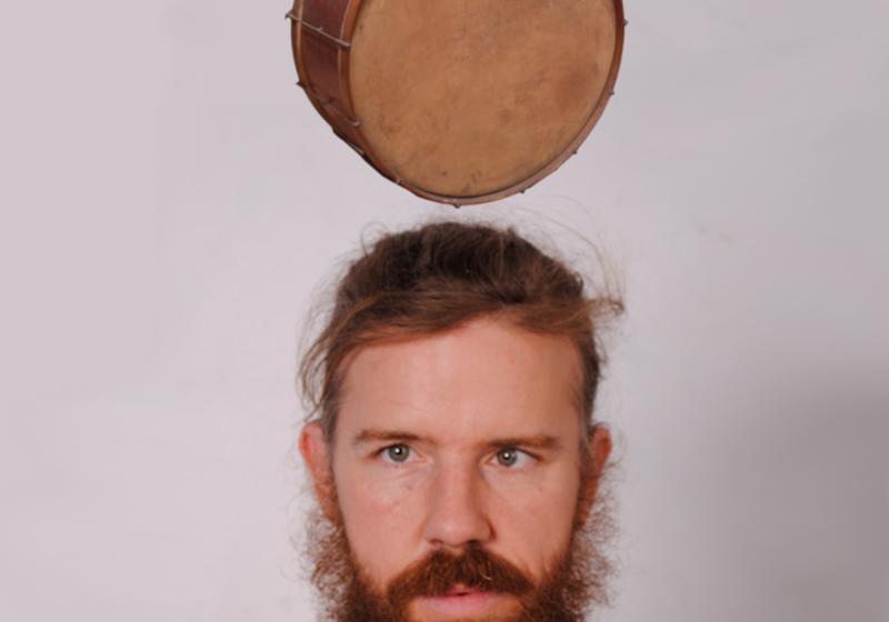 A front-on portrait of Chris O'Connor from the shoulders up. Above Chris' head a drum levitates.