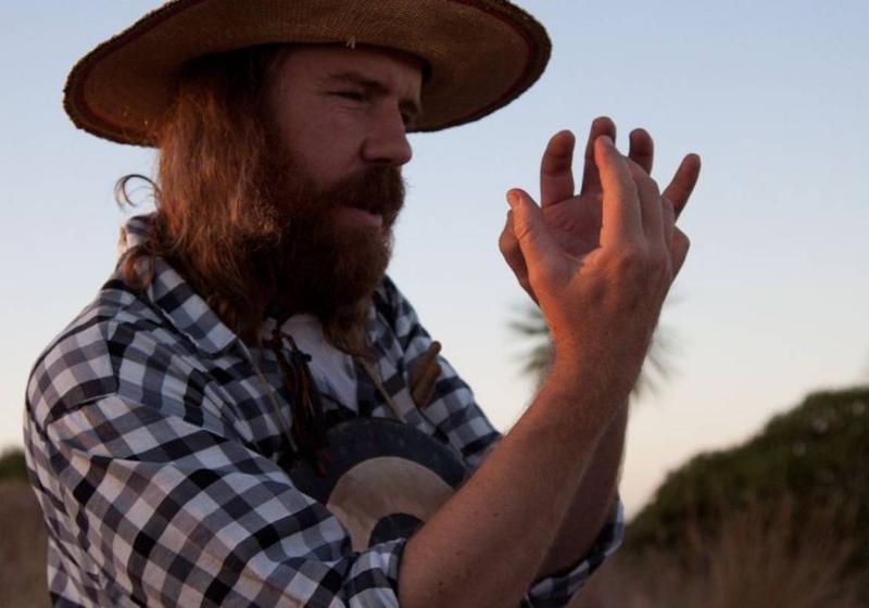 Chris O'Connor stands in front of a sunset explaining something with his hands. He's wearing a large brim hat