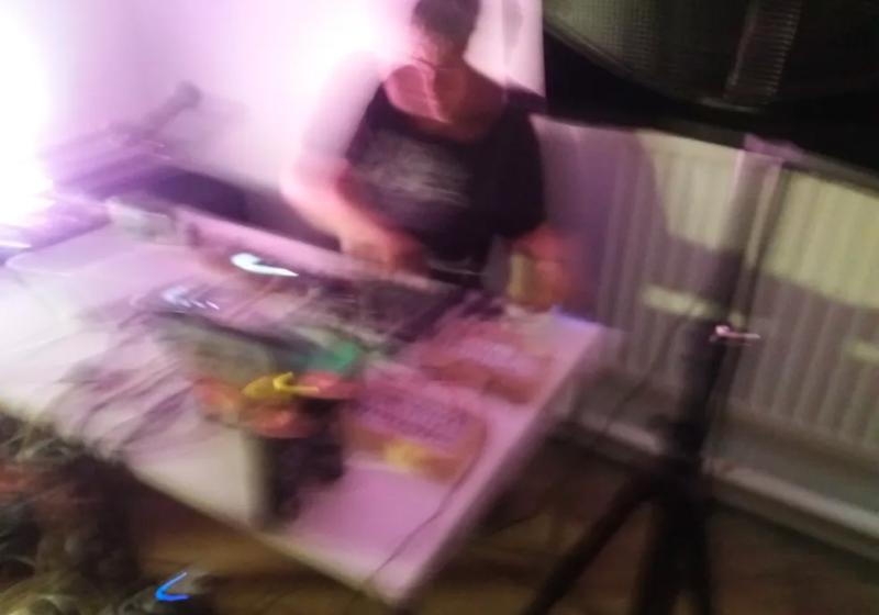 a blury digital camera photo from above an s glass set. S glass has objects arranged across the table including blury electronics. a purple light washes out the left side of the photo. S Glass looks out of breath in an intense moment of performance. 