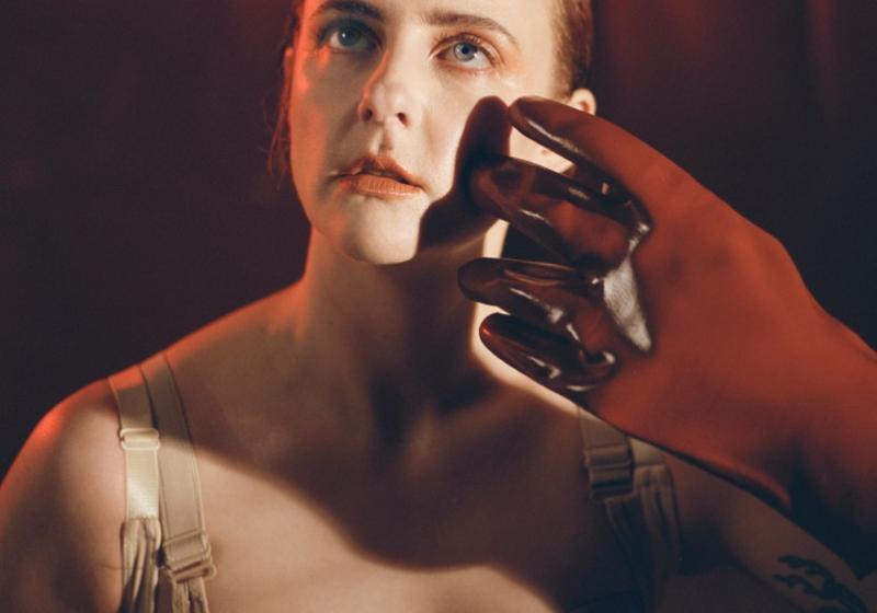 A front-on portrait style photo of i.e. crazy in front of a red curtain background. A red rubber glove reaches out and strokes the side of their face. They are wearing a cream coloured corset and are looking upwards off into the distance, the photo has a dreamy surreal vibe about it. 