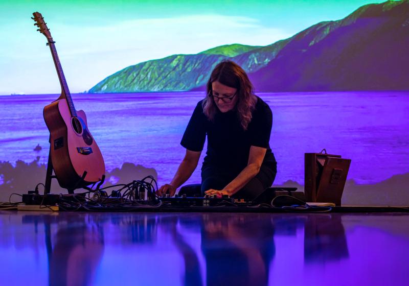 Jacqui O'Reilly sitting down performing in front of a projection of the ocean and a mountain