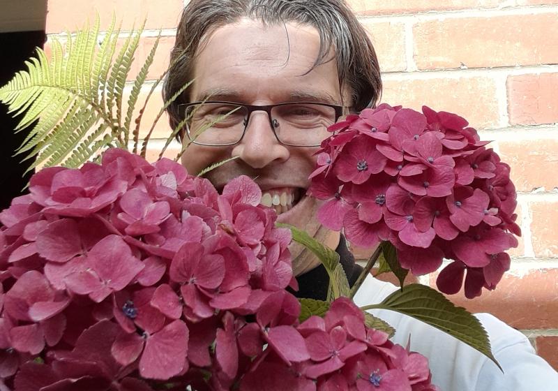 Chris is smiling. He is hiding by surrounding himself with flowers. He is being cheeky