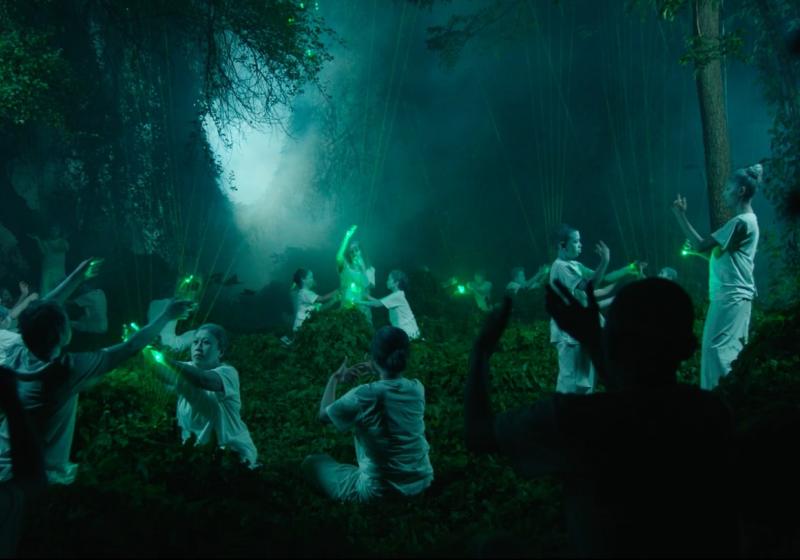 A still from a film by Korakrit, featuring a gathering of 10+ people in a forest touching green lasers/lights beaming