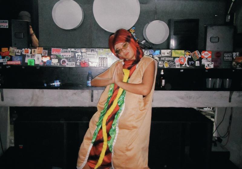 Kristen is dressed as a hotdog in a bun with lettuce and mustard. She is at a nightclub and the walls are covered in stickers
