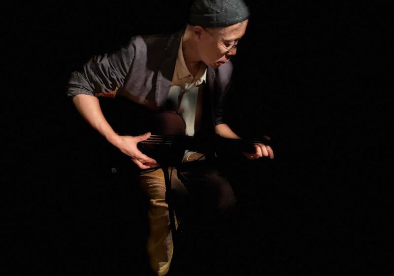 Kawol plays a stringed instrument with a black background