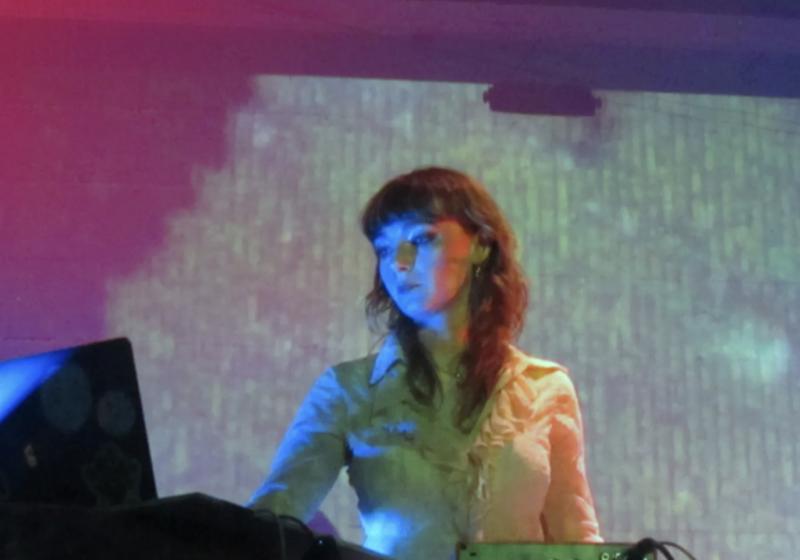 Fox Moss plays live with visuals behind her, looking at her electronic gear as she plays