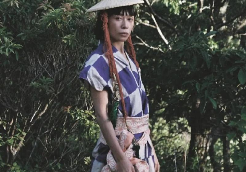 Kina:suttsu stands in front of a bushy backdrop