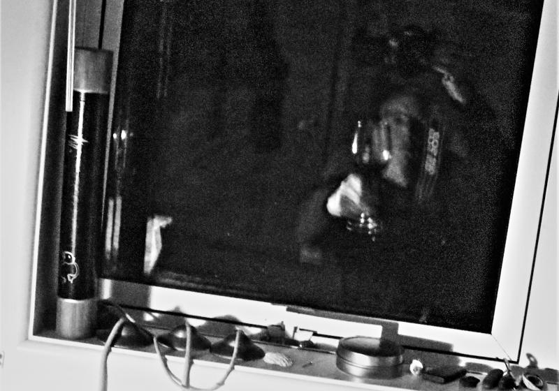 A blurred black and white photo, very grainy. There are chimes on a window ledge. The window is refelcting a figure in motion holding up a glass of red wine in a pose of a toast. The reflection is of the photographer.