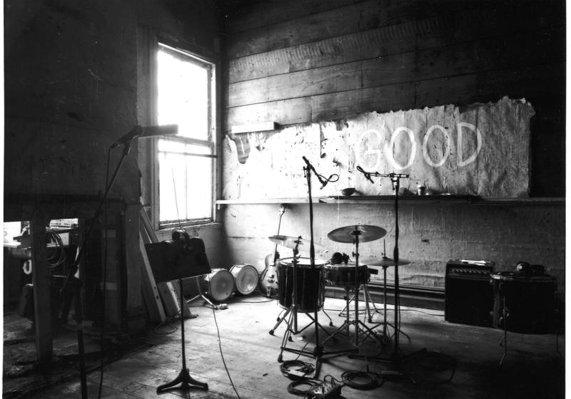 ARC Building Rehearsal Room, black and white photograph of a rehearsal room with one window beaming in bright light into an otherwise very dark room. A drumset, some amps, a music stand and a few mirrors are the only objects in this sparse dingey space.  