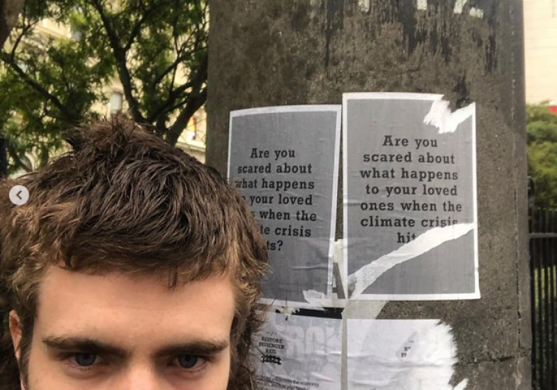 A photo of Baxter in front of a lamp post. The pole has flyers stuck to it, they are slightly torn, and they read "are you ascred about what happens to your loved ones with the climate crisis hits?". Baxter has a mullet and elaborate dangly earrings