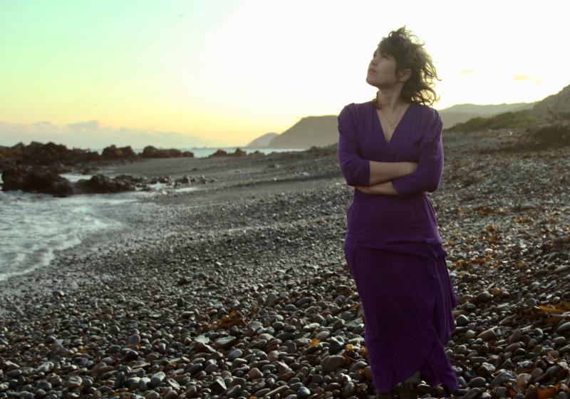 Jess Stands on a rocky beach as the sun is setting. She looks to the sky and has her arms crossed.