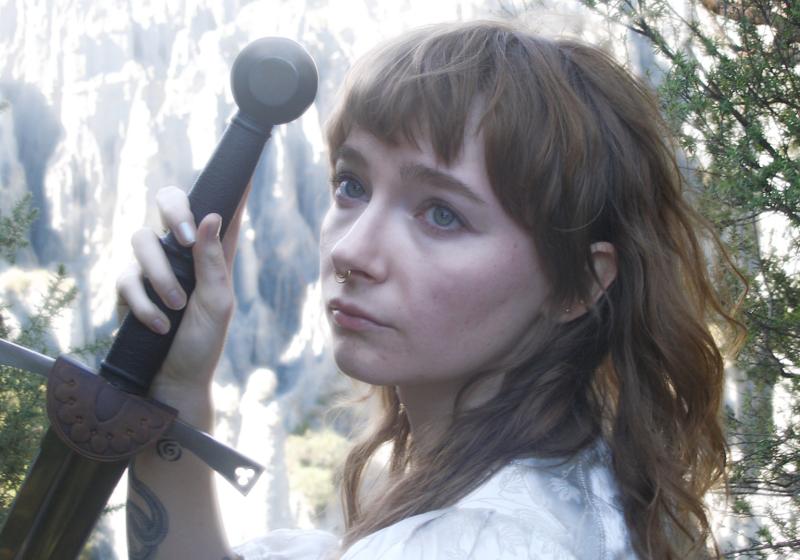A glowy photograph of Fox Moss posing with a sword in front of cliffs looking in the distance. 