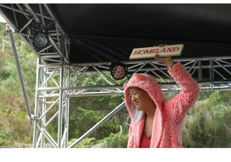 Kristen is on an outdoor stage at Camp a Low Hum. The bush is behind her as she holds up a sign that reads Homeland
