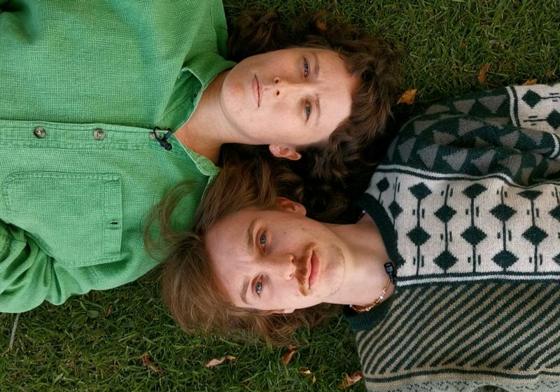 Katie and Oli are lying on the grass lookiing up at the sky. The camera is in the sky looking down at them. Their shoulders are touching and their heads are next to each other. Katie is lying one way and Oli the opposite way.