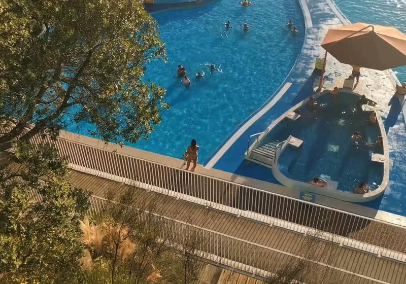 A view down on a pristine blue pool. There are people in the main pool, and also in an inset spa pool. It is clearly a relaxed and beautiful day.