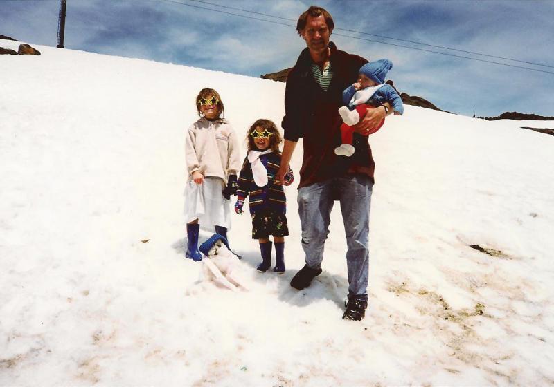 on a mountain of dirty snow stand two children with sunglasses shaped like stars. Their father holds a baby in his arms.