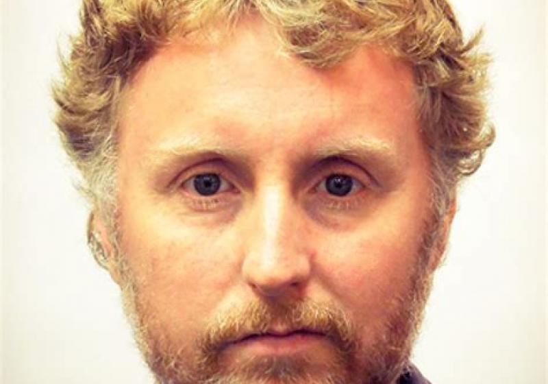 A stark headshot of Mark. He is in front of white wall. This could well be a passport photo.