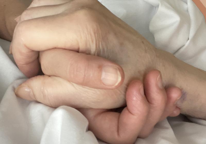 two hands are held. The hand underneath is that of the daughter and composer Chrissie. The hand on top is that of her dying mother. the hands are grasped in love. There is a crumpled sheet that the hand are on, it seems the mother is in bed.