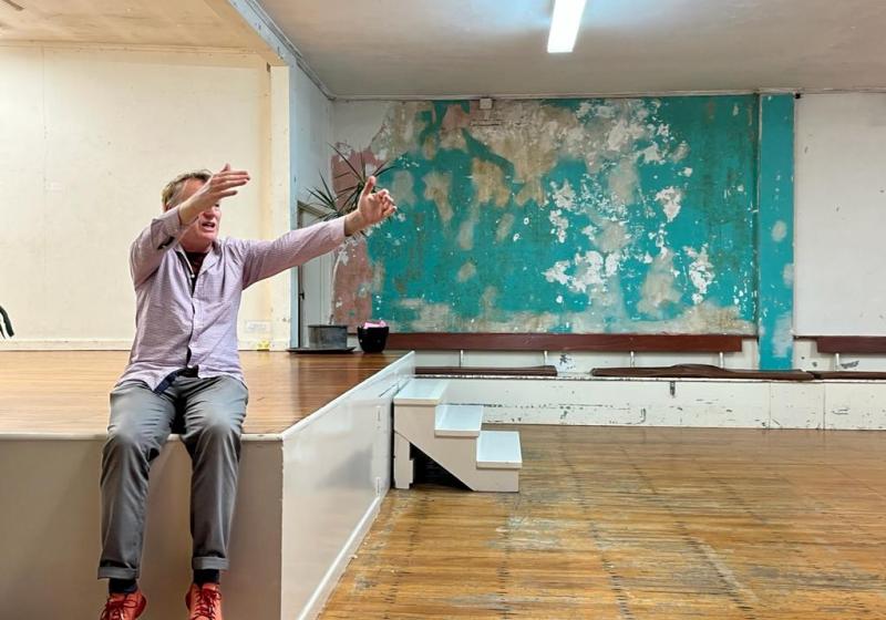 Stephen sits on the corner of a stage at a community hall. The wooden floors are well kept but are starting to show the years of use. The walls are in a state of unkempt life.
