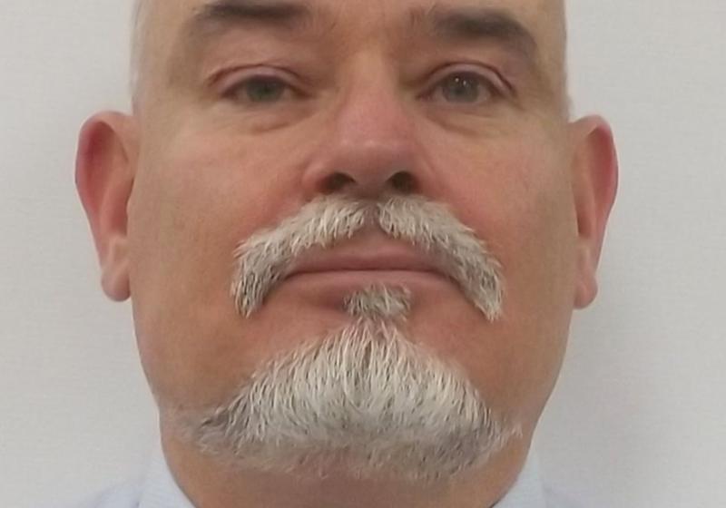 a portrait of Sam. He is looking directly at the camera with a strong and clear face. He has a well groomed goaty and moustache. He is wearing a shirt and tie.