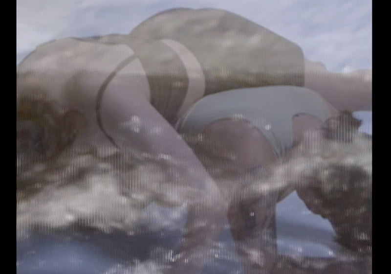 A still from the video work. Two bodies lay over each other cross ways and face down. The bottom person is up on their elbows making for a 'bridge' shape. The image os slightly blurred with a transparent layer that appears to be a churning sea.