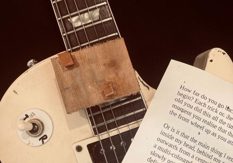 An elctric guitar, Les Paul in style. On top of the strings are the words of Drew's article printed on A4 paper, and a wooden block with rubber squares glued on it.