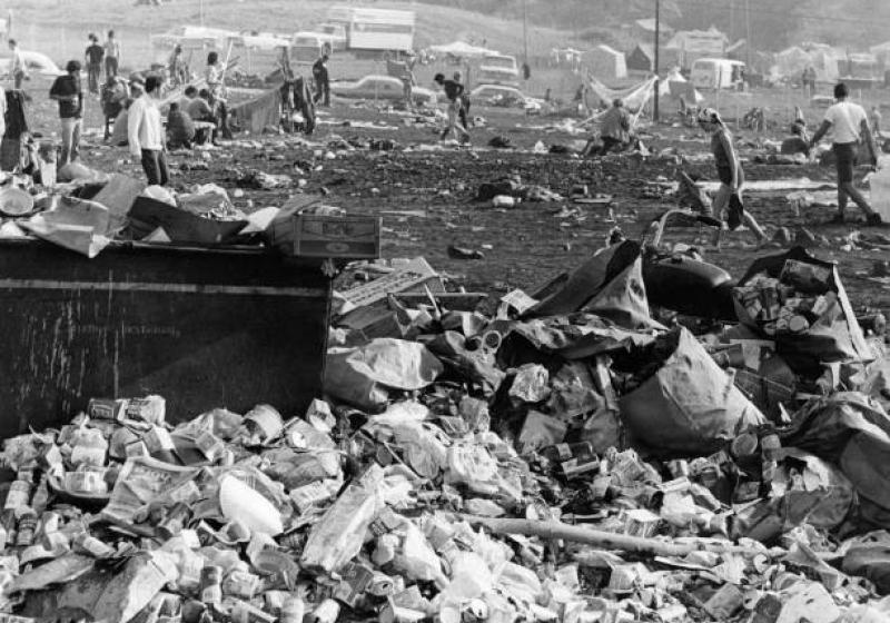 A black and white photo of the afternath of woodstock. There are piles and piles of garbage in what was once a field but is now mud