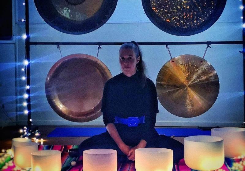 Erika Grant sits cross legged in front of four large gongs that are hanging from a square frame. In front of her there are crystal meditation/prayer bowls that are aglow with warm light