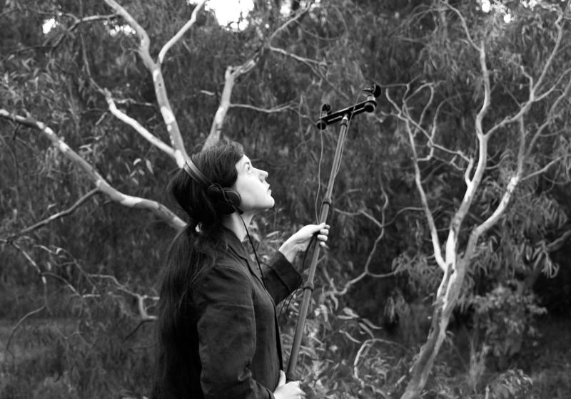 Sally in the bush. She is wearing headphones and has field recording mics on the end of a large boom