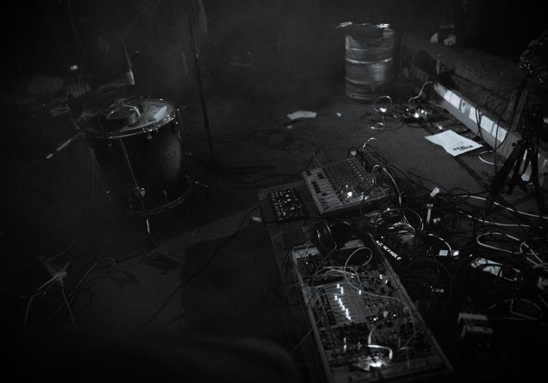 A messy and intriguing set up of cables and boxes and lights. A make shift noise synth. It is in bold black and white. There is a filthy carpet, an empty beer keg, and a dismantled drum kit, in the background.