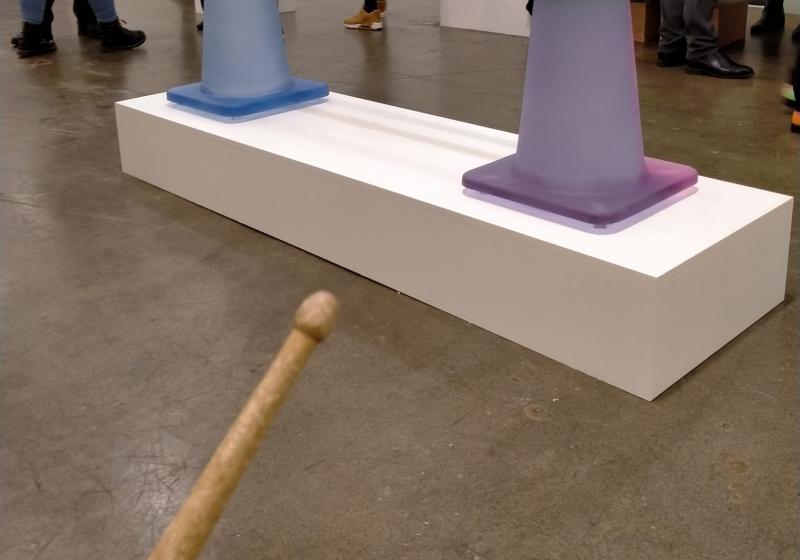 an image from a first person perspective; you are holding a drum stick and advancing towards two lilac coloured roadcones. You are in an art gallery