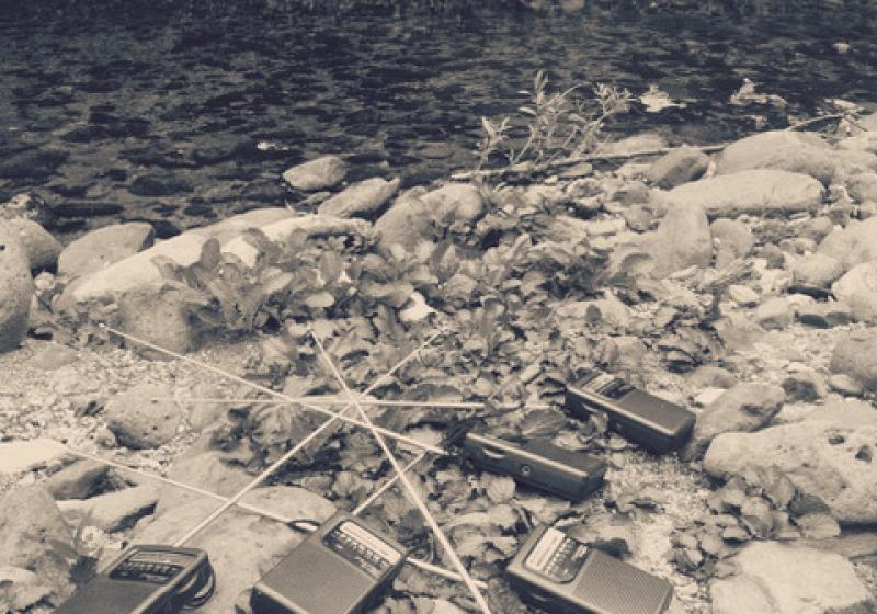 six cheap transistor radios lie around on a river bank. They are scattered amongst the rocks, and their aerials are touching, tangled together. The river flows beside them.