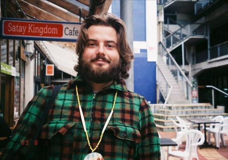 Zak is standing in the left bank off Cuba St. There are signs for restaurants, and outdor tables. Zak is wearing a warm bush shirt, and has a thick beard. He smiles gently through his whiskers.