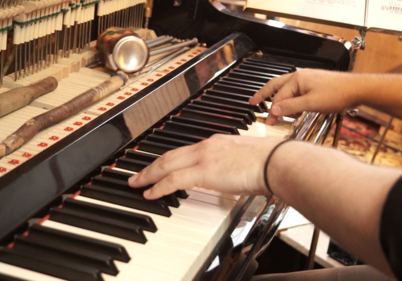 an upright piano with the cover off for access to the strings and hammers. Some hands are playing the piano keys. there are various objects leaning on the mechnical structures, a can, some pices of wood, some lids...