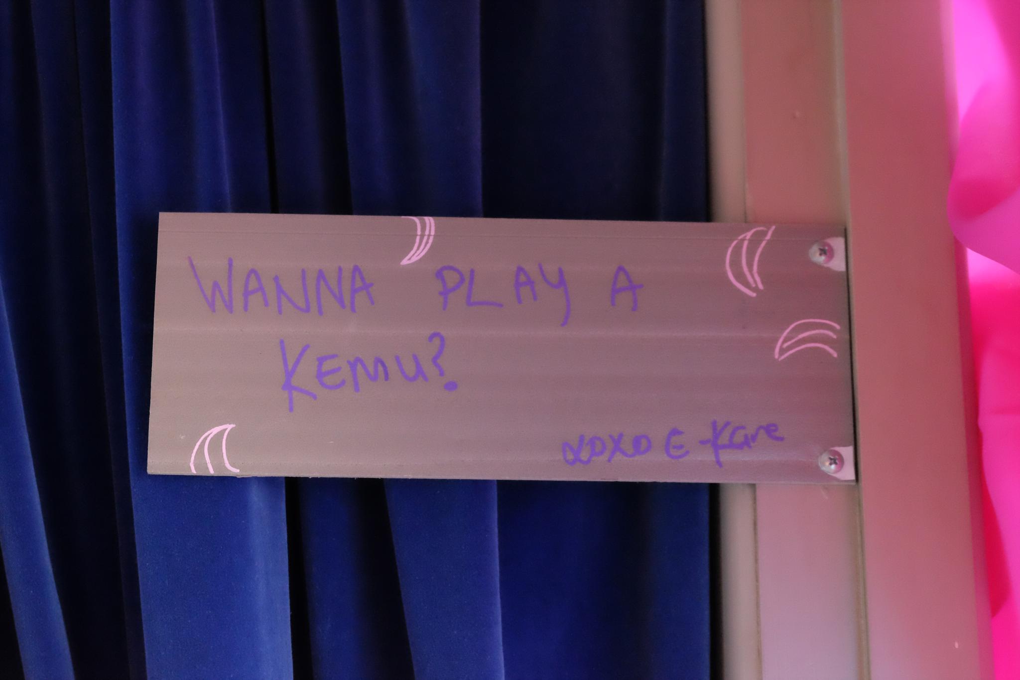 Paint pen writing on a plastic board that reads "Wanna play a kemu? XOXO E-Kare". The sign points visitors into the second gallery room.