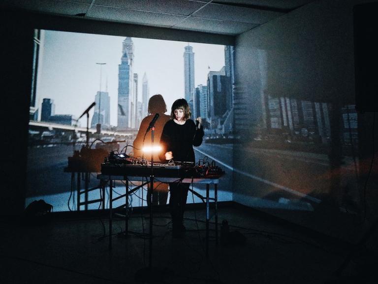 Jess Playing live, an array of electronics in front of her, and a city scene projected on the wall behind her.