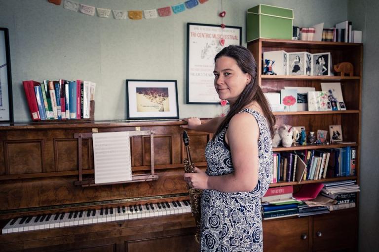 Jasmine standing in front of an an old upright piano. It is a very doemnstic setting with photos on top of the piano and a pleasingly unitdy bookshelf in the background.