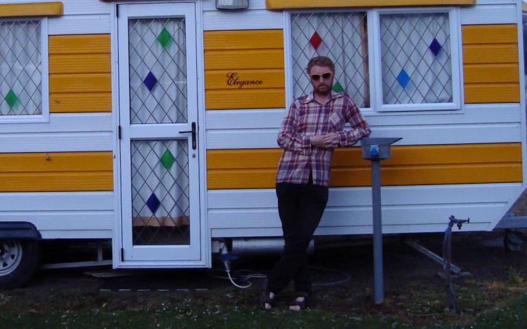 Mark is leaning nonchanontly in front of a happy yellow and white caravan