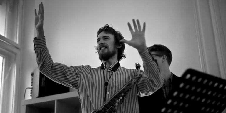 A back and white photo of Jake holding his hands up in a gesture of conducting some musicians unseen. There is a music stand to one side of him.