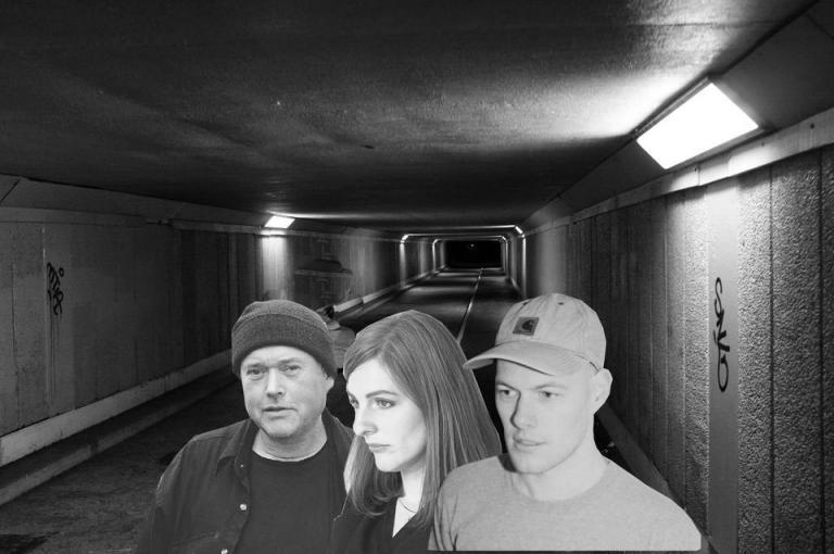 A black and white photo of the pedestrian tunnel under Wellington Airport. Collaged on top are the head and shoulders of Amy, Cory, and Omit.