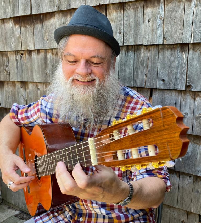 Chris has a large grey beard and is wearing a small pork pie hat. He is playing a nylon string guitar and is smiling with a large joyful smile.