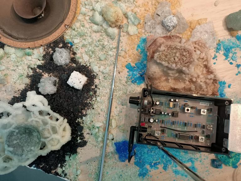 A table with whote a blue (copper?) crystals growing on it. There is a raw speaker cone and an open electronic device.